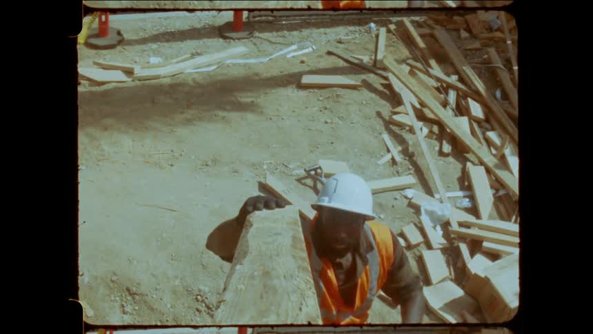 A construction worker in a hard hat receives a plank amid scattered lumber