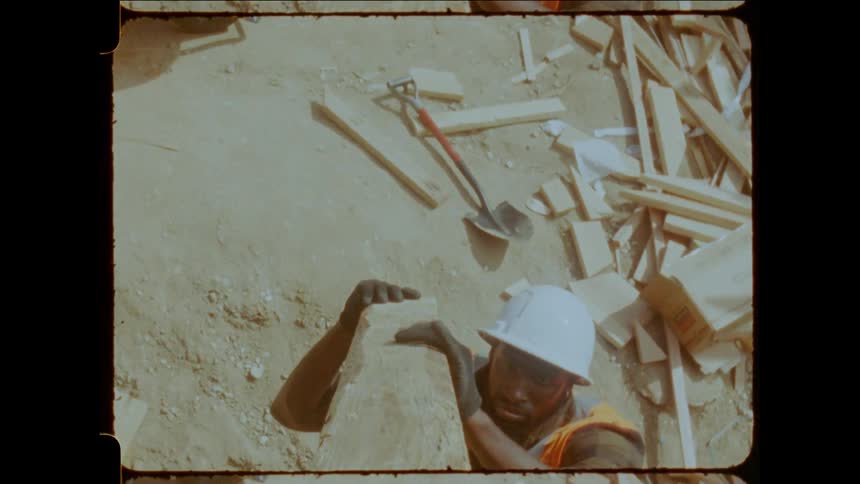 A wooden beam is passed from worker to worker both wearing hard hats and vests at a construction site