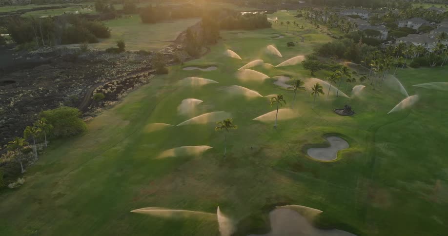 A golf course is being irrigated by sprinklers with palm trees and buildings visible in the background