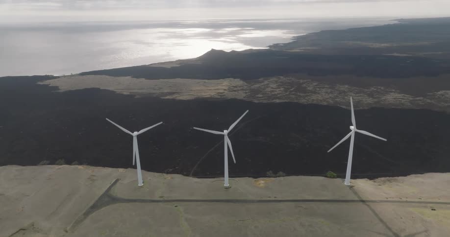 A series of windmills stand on a hill near a cliff rotating with an ocean view on the other side of the cliff at midday