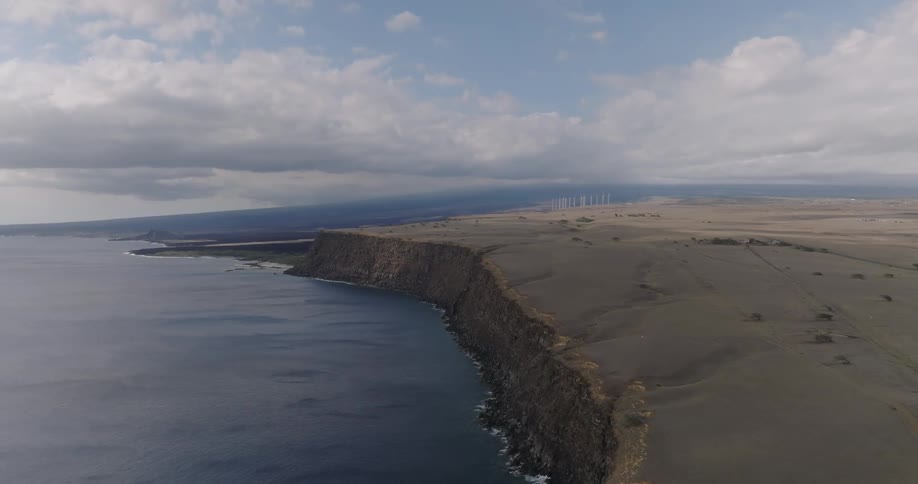 An aerial view shows a stark coastal landscape where a dark cliff edge meets the ocean with wind turbines visible in the distance