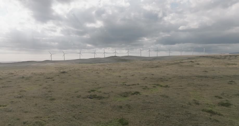 A series of wind turbines rotate in the distance beyond a grassland on a cloudy day