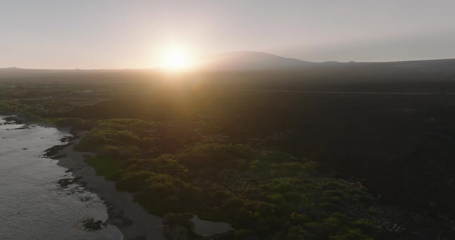 An aerial view of a coast with various green vegetation next to the sea on a day with the sun rising on the horizon