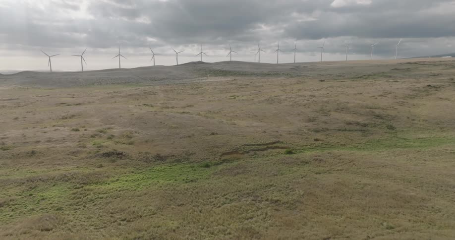 A wide aerial shot of a wind farm with multiple wind turbines on rolling plains under a cloudy sky