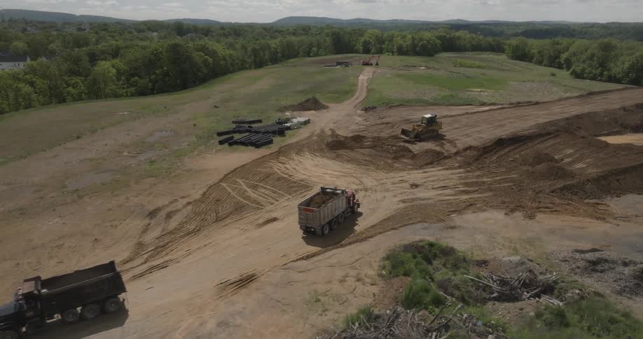 A truck drives past another truck heading toward an excavator carrying sand as the excavator spreads sand in an area surrounded by vegetation at midday