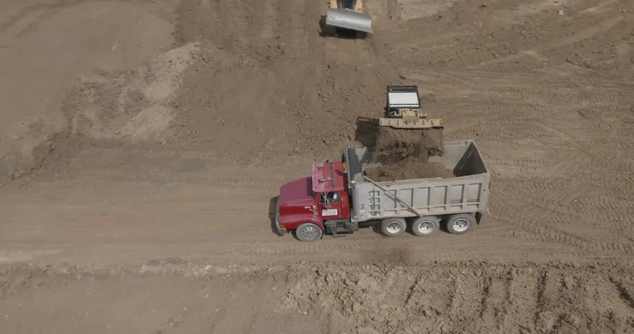 A skid loader loading sand in a dump truck while a bull dozer pushes sand forward