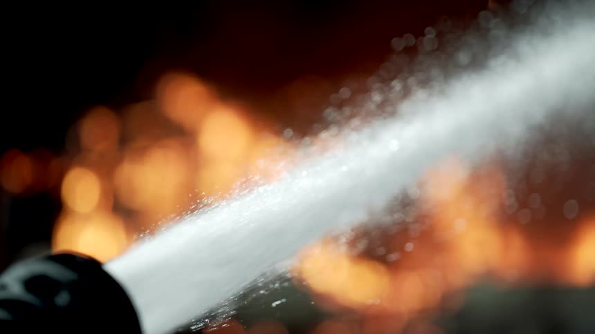 A firefighters hose releases water toward a burning building to extinguish the fire