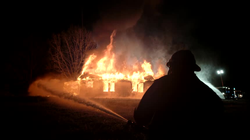 Fireman silhouette hosing down a house engulfed in flames at night