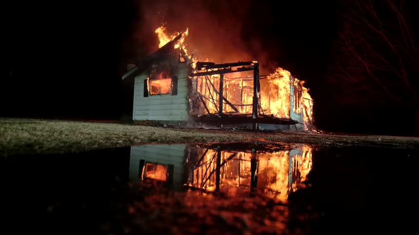 Remainder of house engulfed in flames reflecting off the water near shoreline at night