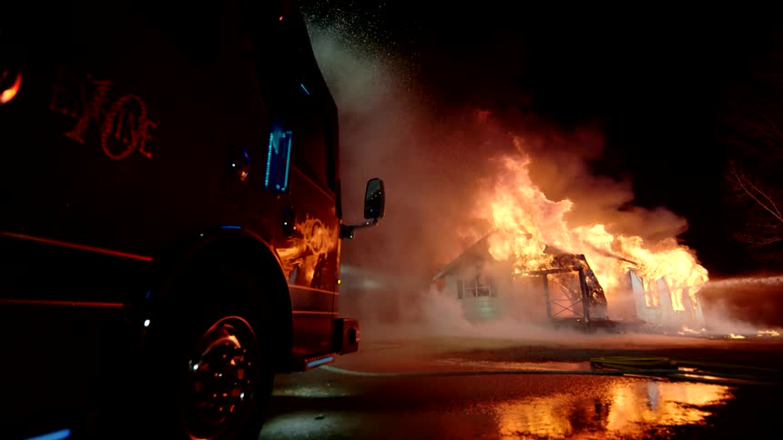 Vehicle drips water while a firefighting jet of water hits a house fire at night