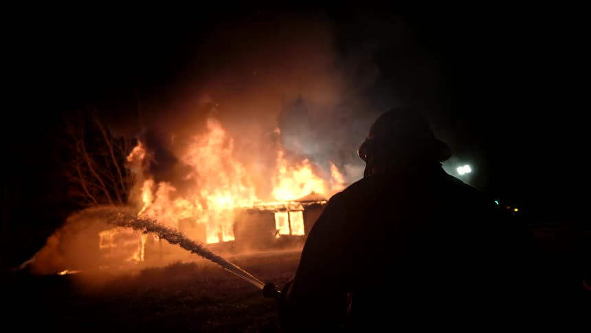 The firefighter is spraying water on the house that is up in flames