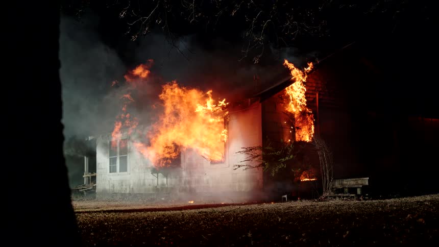 A house is on fire with fire coming out of the windows there is smoke coming from the fire and the video is eerie because it is being shot at night