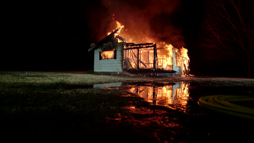 A house is completely engulfed in flames at night reflecting off a wet surface nearby