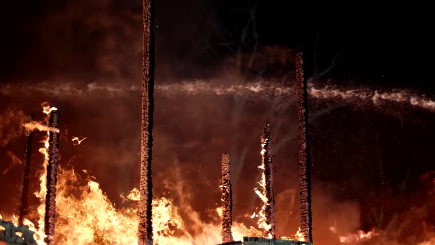 Intense flames engulf the remains of a burnt structure while water is sprayed onto the fire
