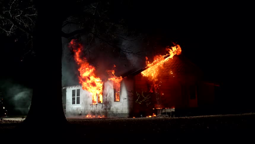 A wooden house that is on fire with high orange flames on a dark night
