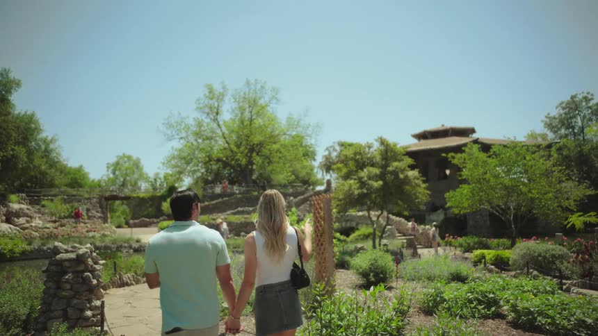 A couple walks hand in hand along a stone pathway in a lush garden surrounded by vibrant flowers trees a pond and a rustic building in the background