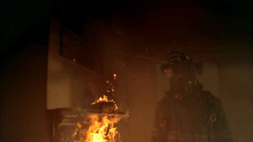 Firefighters walk through a smoky building past a burning piece of furniture