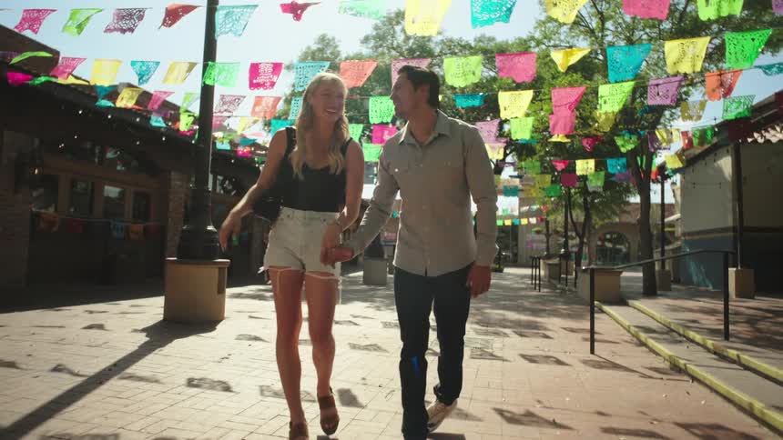 A couple is holding hands and standing close together under a canopy of colorful papel picado banners in a sunlit festive outdoor plaza
