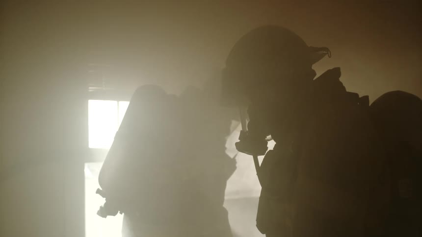 Two firefighters in protective gear move through smoke