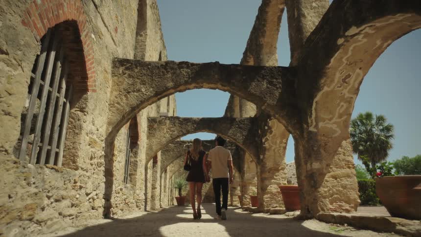 Couple holding hands strolling though outdoor corridor of a stone mission