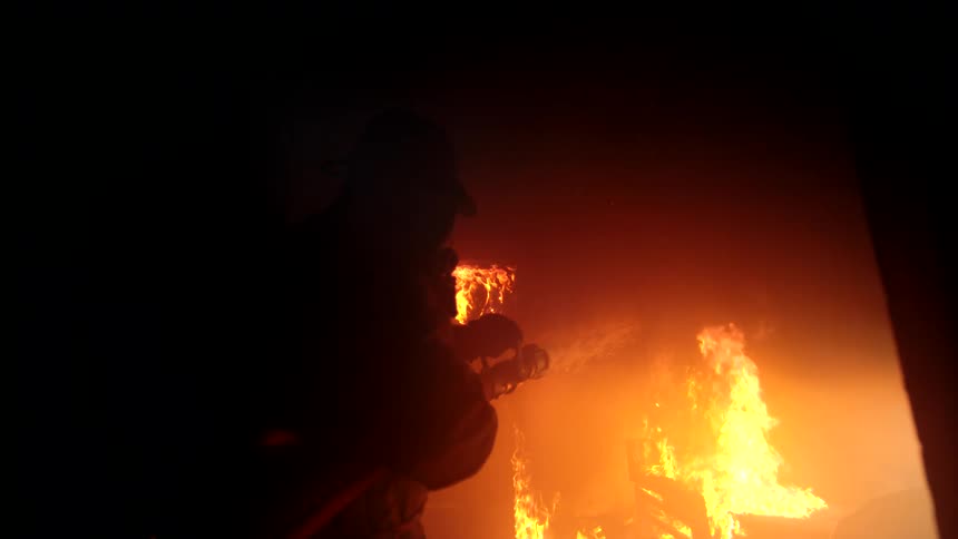 A firefighter in full gear is spraying water onto a burning structure at night