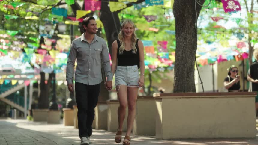 A couple of a man and a woman holding hands walking on a sidewalk with several colorful decorative flags above them