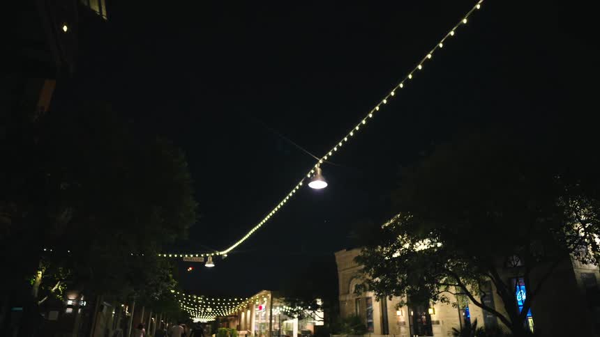 A brightly lit street at night features people walking near buildings adorned with strings of lights