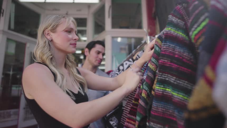 Woman admiring dresses and mexican hoodies at an outdoor market as male walks behind her