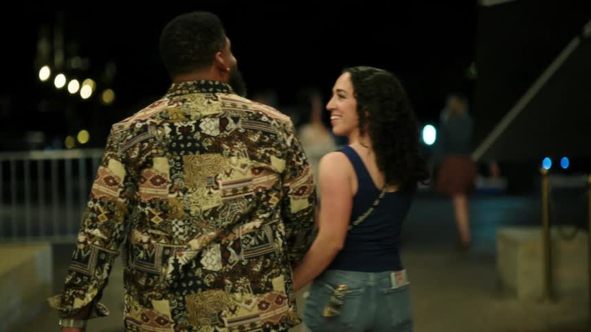 A man in a patterned shirt and a woman in denim shorts walk hand in hand down a city street at night