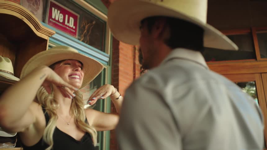 A woman putting a cowboy hat on her head and then smiling at a man and she is spinning around holding his hand