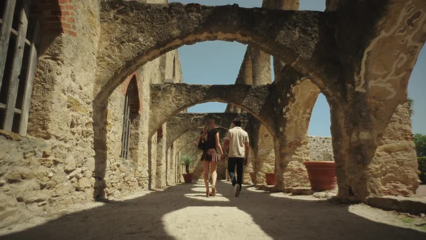 A couple hold hands while they walk under the stone arches