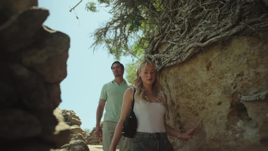 A man and woman are walking on a rocky path beside a stone wall covered in roots under a bright sky
