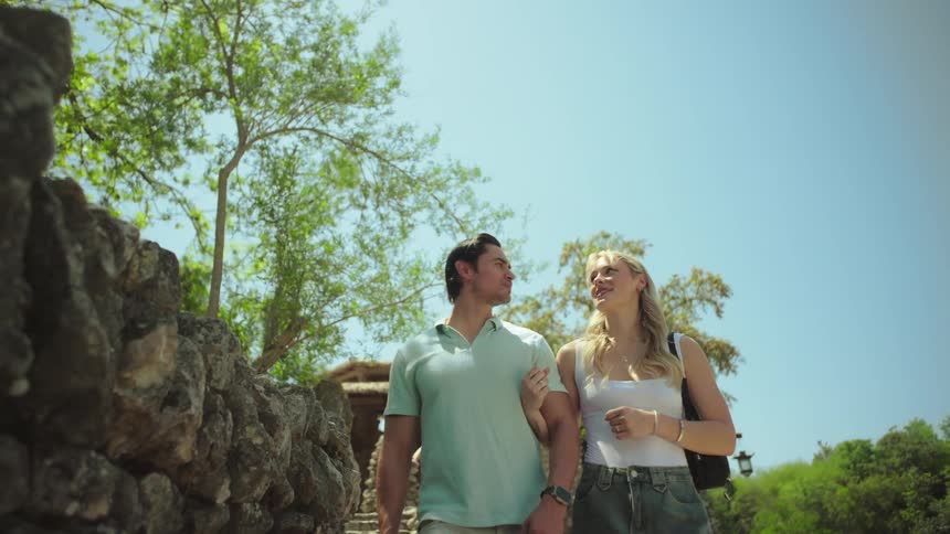 A man and woman are walking arminarm along a path with rocky walls and lush foliage under a bright sky