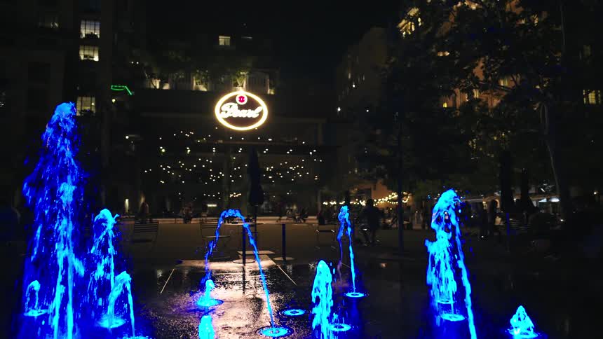 People in outdoor restaurants at night and a fountain with blue led light in the foreground