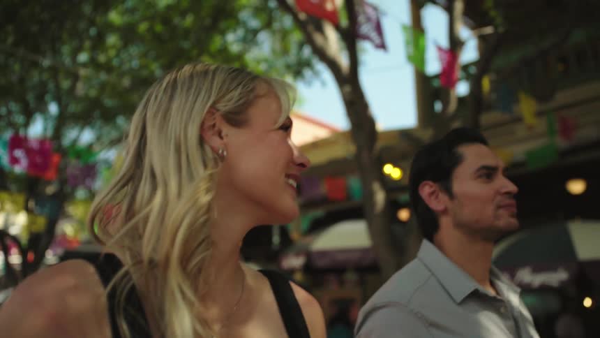 Couple smiles while walking along a street lined by shops and cafes on a summer day