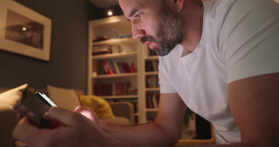 Closeup of a young man with a beard holding a credit card while making a purchase on his phone in a living room