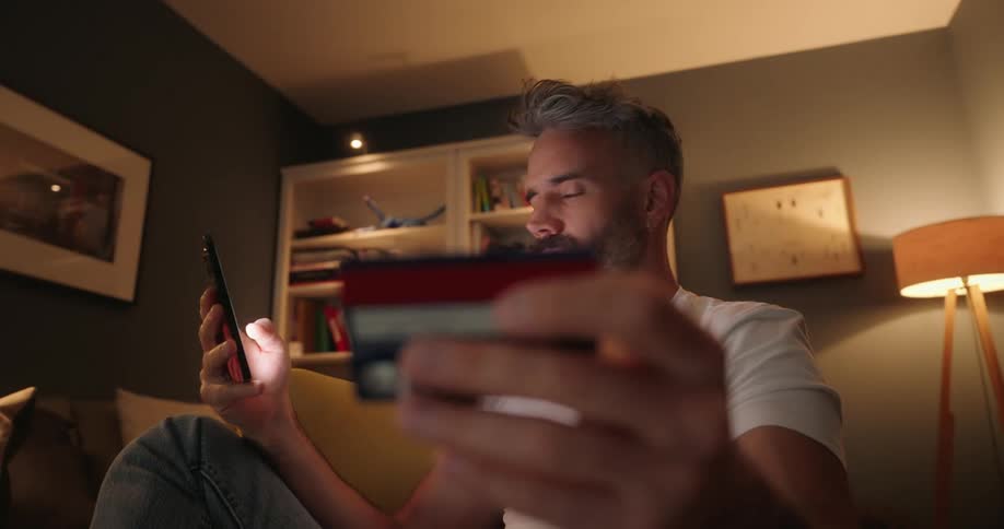 Man sitting on a chair in the living room using his phone and looking at information on a credit card