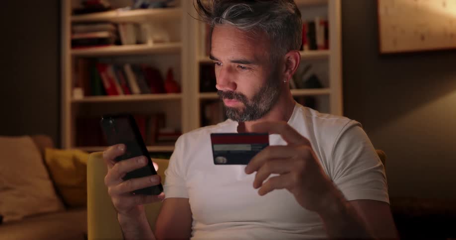 A man sitting on a chair in the living room using his phone and holding a credit card