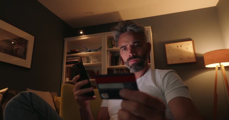 A man sits indoors holding a credit card in one hand and a smartphone in the other seemingly engaged in an online transaction