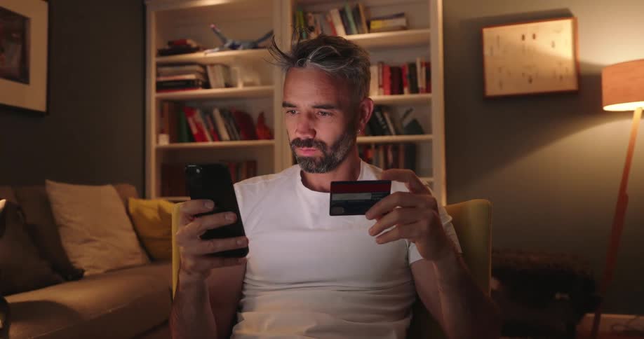 A man sitting on a chair indoor types on a mobile phone his credit card details