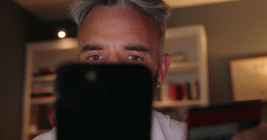 A man closely examines his smartphone while entering credit card details in a dimly lit room