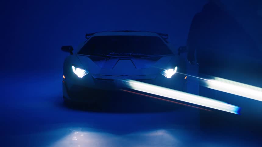 A man walks toward a lamborghini parked in a garage after turning on the blue garage light and the lamborghinis headlights come on