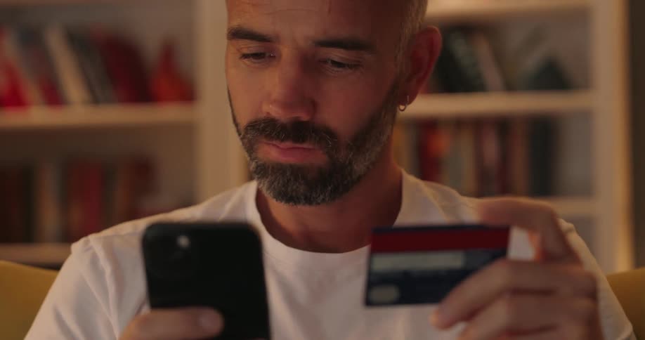 A older man with and salt and facial hair looks back and forth between his credit card and cell phone he is sitting in a chair with a white book case full of books behimd him