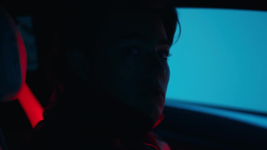 A man sitting in the passenger seat of a car rolls down the window with flickering blue light visible outside while the inside of the car remains dark