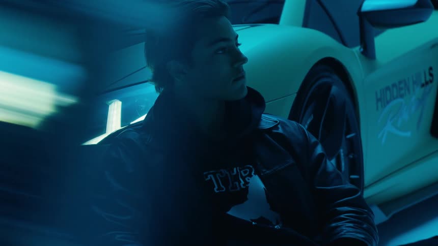 A young man sitting on the ground leaning against a sports car posing for a photo shoot