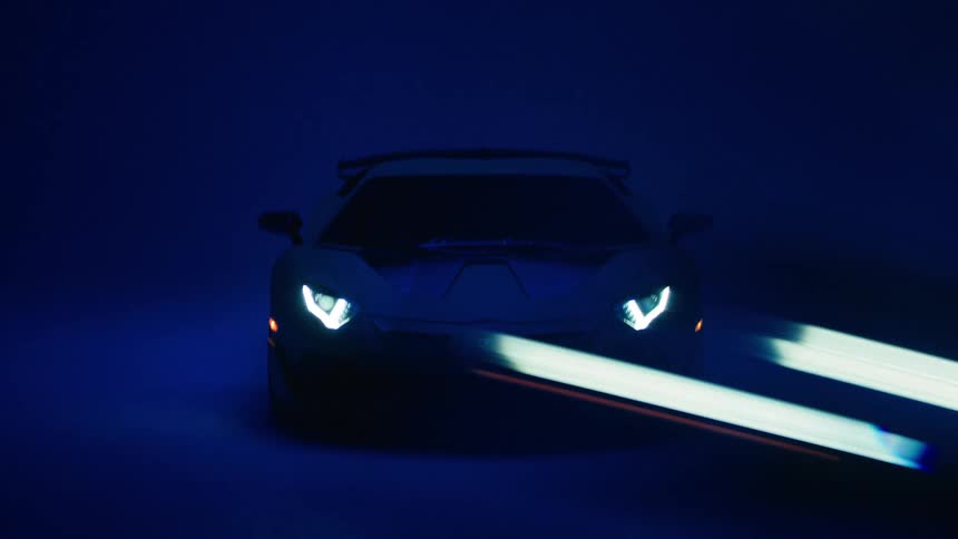 A man walks toward a lamborghini parked in a garage with its headlights on and the blue garage light illuminating the space at night