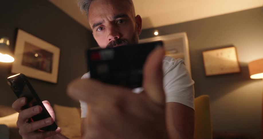 A man sits indoors holding a phone and entering credit card details with a focused expression