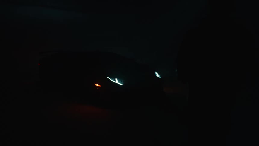 A sports cars headlight lights up in a dark garage as the garage light flickers and a man walks beside the car