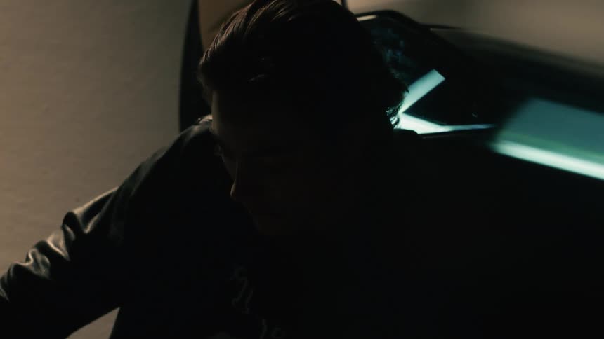 A man sits in front of a sports car in a garage at night with the cars headlights turned on