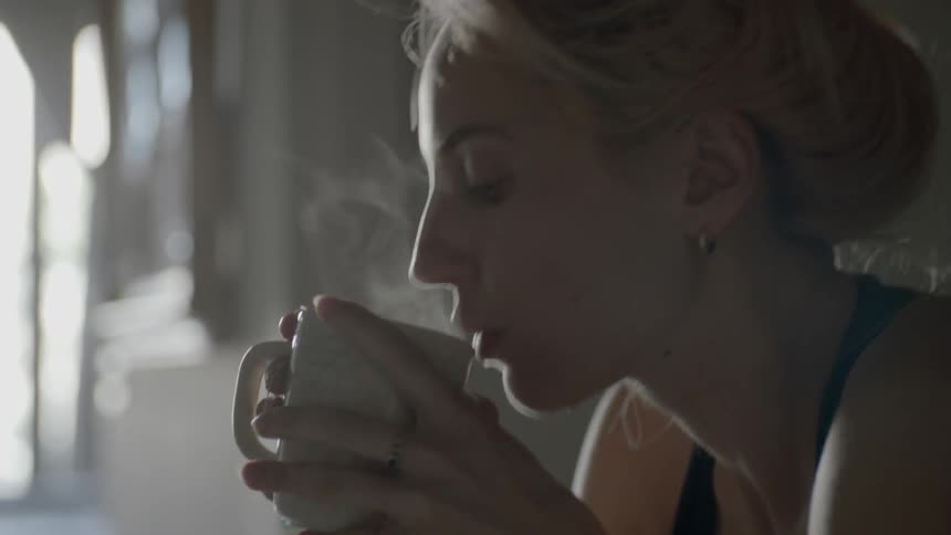A woman holding a hot cup of coffee in the kitchen and blowing on it